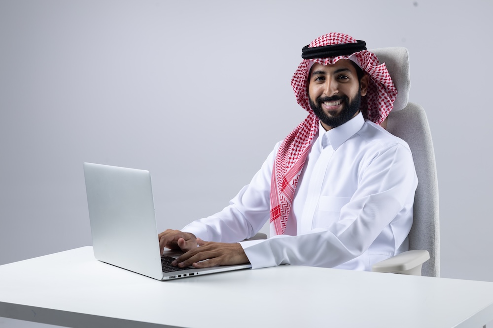مبرمج سعودي يرتدي الثوب والشماغ يجلس على مكتب أبيض أمام جهاز لابتوب ويبتسم أثناء عمله عن بُعد